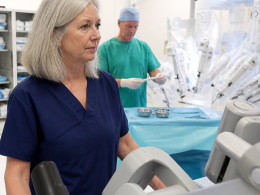 Woman in navy scrubs observing robotic surgery with concerned surgeon adjusting gloves nearby in an operating room.