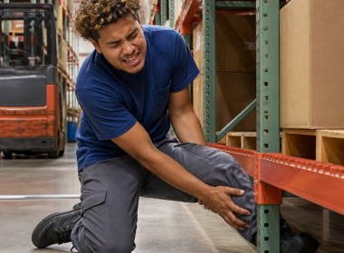 Man trapped under a warehouse rack, clutching his crushed lower leg with a pained expression.