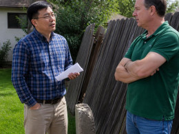 Man with glasses holding a paper standing by old fence talking to neighbor crossed arms