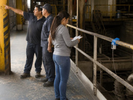 Latina woman inspecting taped sensor on factory railing while two men discuss missing guardrails nearby.