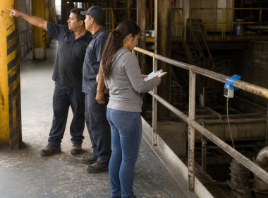 Latina woman inspecting taped sensor on factory railing while two men discuss missing guardrails nearby.