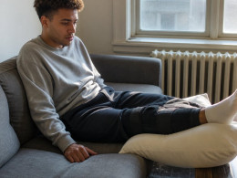 Young man with knee brace sitting on couch, looking worried while holding phone beside him.