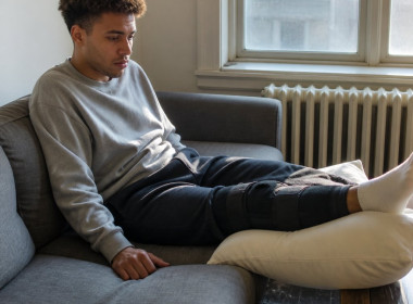 Young man with knee brace sitting on couch, looking worried while holding phone beside him.