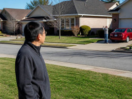 Man on lawn watching neighbor filming rooftop solar panels from driveway.