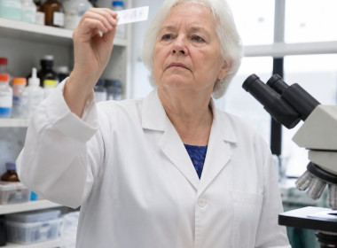 Older white woman in lab coat inspecting stained slide in pathology lab