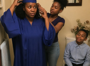 Woman adjusting graduation cap in bathroom mirror while her daughter helps and son watches