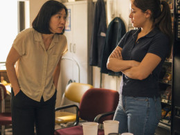 Maya speaking quietly to Danielle in a cluttered break room, Danielle looking detached with crossed arms.