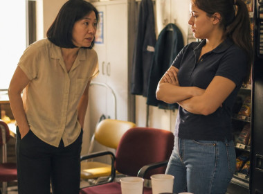 Maya speaking quietly to Danielle in a cluttered break room, Danielle looking detached with crossed arms.