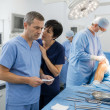 Nurse holding a consent form discussing quietly with charge nurse while a surgeon preps a patient’s left knee in the OR.