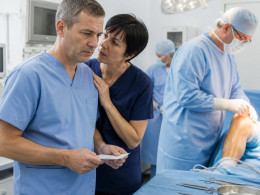 Nurse holding a consent form discussing quietly with charge nurse while a surgeon preps a patient’s left knee in the OR.