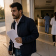 Man leaving hospital holding documents with a serious expression, hospital staff blurred in background.