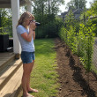 Woman on back porch photographing a row of young saplings along a fence line in a suburban backyard.
