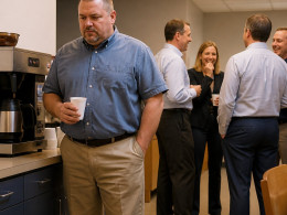 Man near coffee machine in an office break room, coworkers nearby but distant, fluorescent lights overhead.