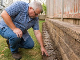 Man inspecting damp soil line by fence near retaining wall in backyard.