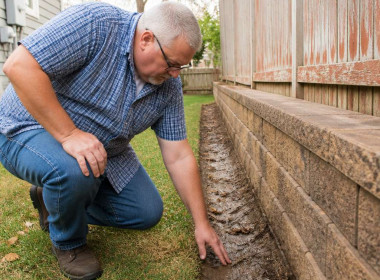 Man inspecting damp soil line by fence near retaining wall in backyard.