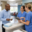 Man speaking to two nurses at a surgery center reception desk, both nurses showing confusion while discussing paperwork.