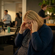 Woman holding her head in a quiet office corner, isolated from noisy coworkers.