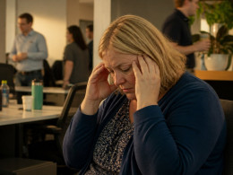 Woman holding her head in a quiet office corner, isolated from noisy coworkers.