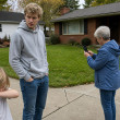 Young man and older woman near cracked sidewalk, woman filming seam, suburban front yard.