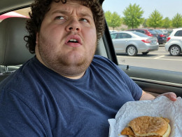 Man in car holding a sad-looking fast food sandwich while looking up in disbelief