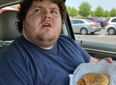 Man in car holding a sad-looking fast food sandwich while looking up in disbelief