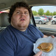 Man in car holding a sad-looking fast food sandwich while looking up in disbelief