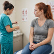 Freckled young woman watching a medical assistant dip a urine test cup in a clinic room.