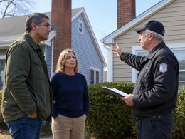 A man and woman stand near their homes' chimneys while a uniformed inspector gestures at the brick stack on the property line.