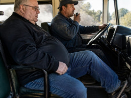 Man seated on bus pressing brake pedal, looking concerned; driver wearing blue cap speaks into radio inside smoky bus.