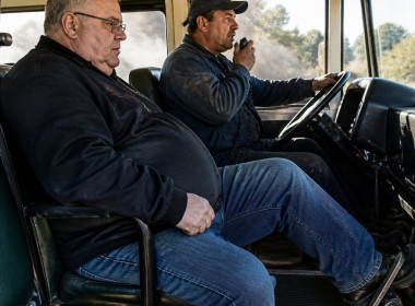 Man seated on bus pressing brake pedal, looking concerned; driver wearing blue cap speaks into radio inside smoky bus.