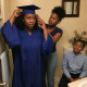 Woman adjusting graduation cap in bathroom mirror while her daughter helps and son watches