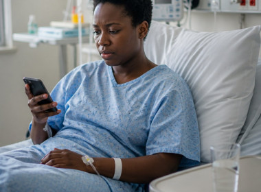 Black woman in hospital bed looking at her phone with a confused expression.