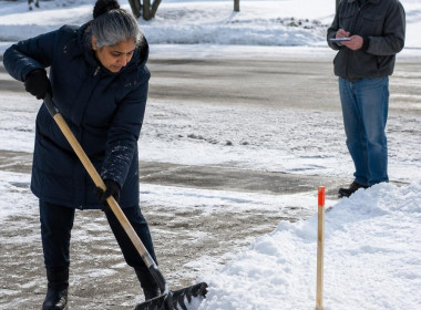 Woman shoveling snow on driveway while neighbor watches and takes notes across the street
