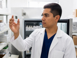 Young Latino man in lab coat examining data logger beside vaccine refrigerator in clinic supply room.