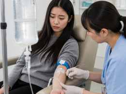 East Asian woman watching a cloudy saline bag in an IV lounge while a nurse adjusts her IV.