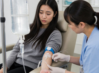 East Asian woman watching a cloudy saline bag in an IV lounge while a nurse adjusts her IV.