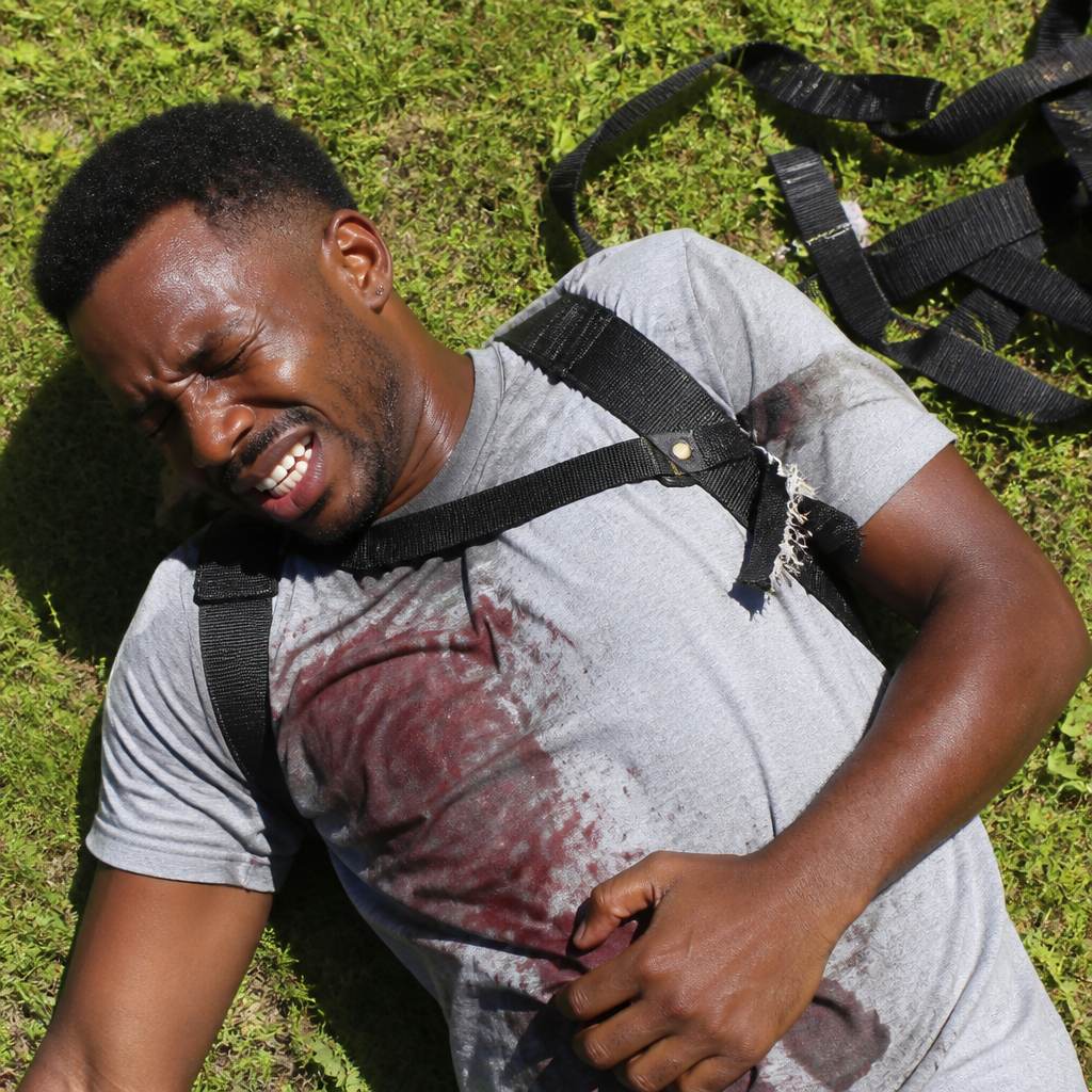 Man lying on grass in pain with broken harness strap showing loose stitches.